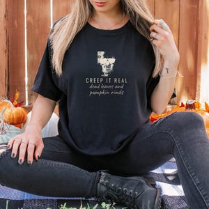 Black Comfort Colors t-shirt printed with the words Creep It Real Dead Leaves And Pumpkin Rinds and a drawing of a grinning skull with black sunglasses, worn by a woman