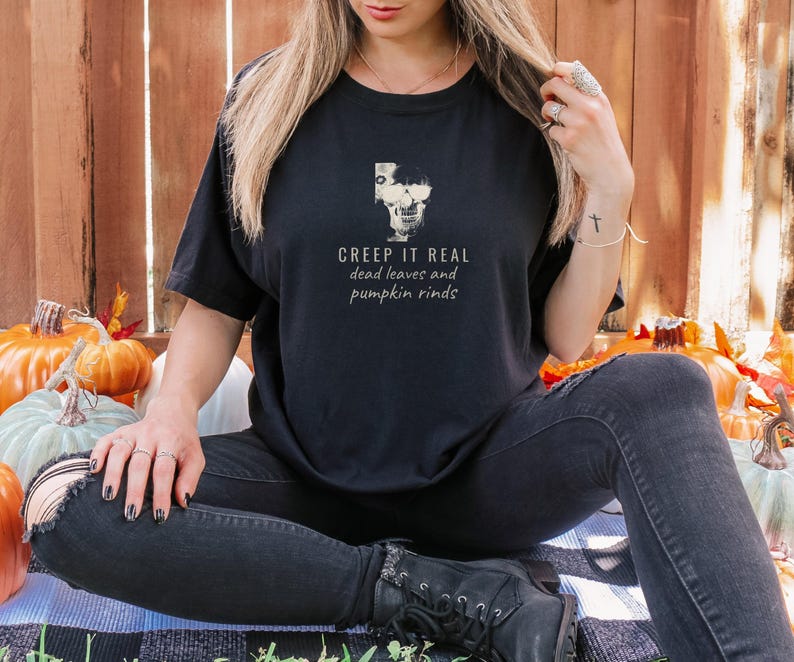 Black Comfort Colors t-shirt printed with the words Creep It Real Dead Leaves And Pumpkin Rinds and a drawing of a grinning skull with black sunglasses, worn by a woman