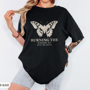 Black Comfort Colors t-shirt printed with the words Burning The Midnight Oil With The Moths and a drawing of a cecropia moth with feathery antennae, worn by a woman