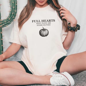 Ivory Comfort Colors t-shirt printed with the words Full Hearts Heavy Plates And Second Helpings and a drawing of a pumpkin surrounded by fall leaves and small acorns, worn by a woman