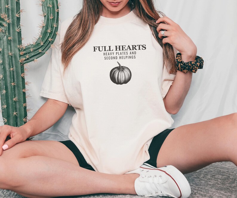 Ivory Comfort Colors t-shirt printed with the words Full Hearts Heavy Plates And Second Helpings and a drawing of a pumpkin surrounded by fall leaves and small acorns, worn by a woman