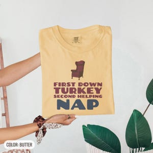Butter Comfort Colors t-shirt printed with the words First Down Turkey Second Helping Nap and a drawing of a vintage brown recliner with a footrest extended outward, folded flat lay