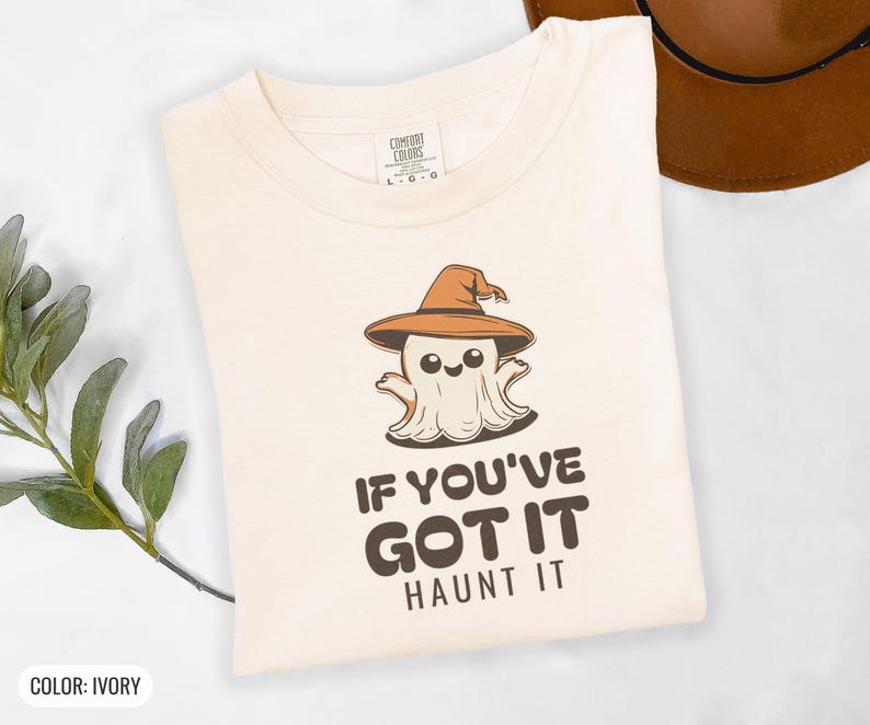 Ivory Comfort Colors t-shirt printed with the words If You'Ve Got It Haunt It and a drawing of a cute retro cartoon ghost wearing a witch hat, folded flat lay