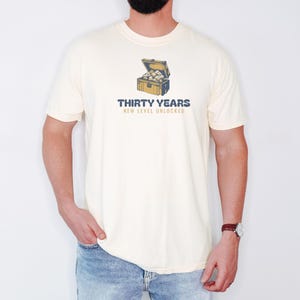 Ivory Comfort Colors t-shirt printed with the words Thirty Years New Level Unlocked and a drawing of a pixel-art treasure chest bursting open with light, worn by a man