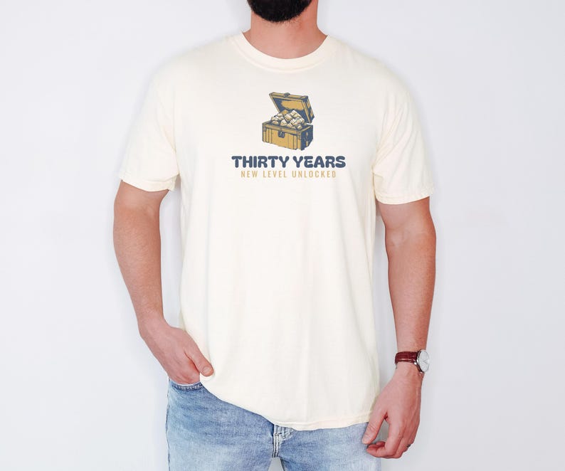Ivory Comfort Colors t-shirt printed with the words Thirty Years New Level Unlocked and a drawing of a pixel-art treasure chest bursting open with light, worn by a man