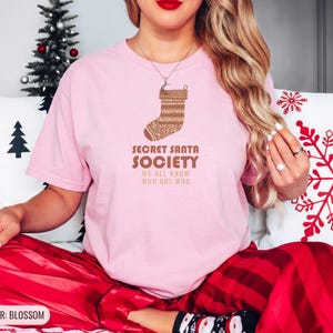 Blossom Comfort Colors t-shirt printed with the words Secret Santa Society We All Know Who Got Who and a drawing of a knitted wool stocking stuffed with stick candy, worn by a woman