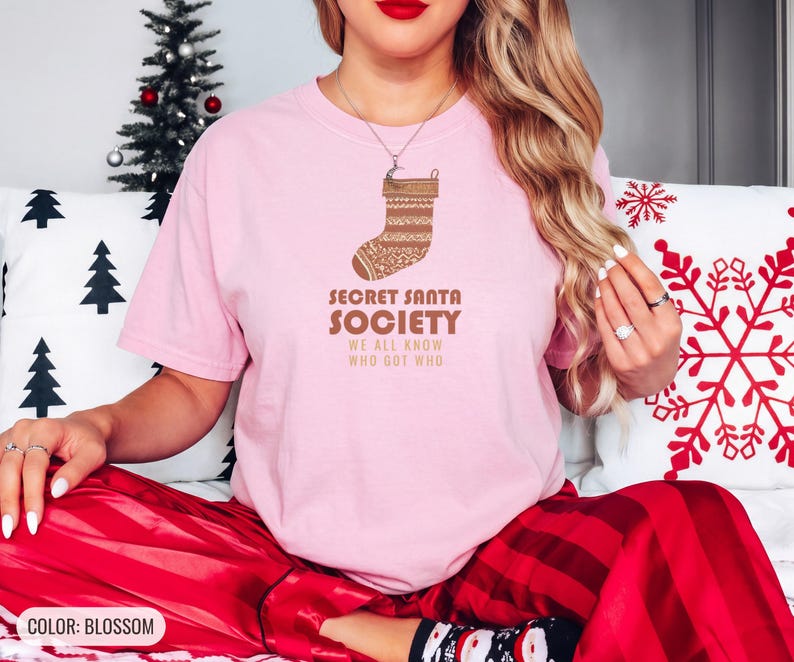 Blossom Comfort Colors t-shirt printed with the words Secret Santa Society We All Know Who Got Who and a drawing of a knitted wool stocking stuffed with stick candy, worn by a woman