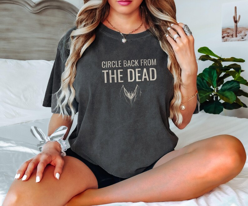 Pepper Comfort Colors t-shirt printed with the words Circle Back From The Dead and a drawing of a vintage silver paperclip bent into the shape of a bat, worn by a woman