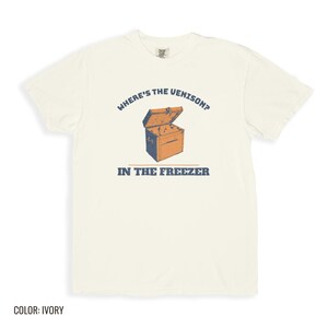 Ivory Comfort Colors t-shirt printed with the words Where'S The Venison? In The Freezer and a drawing of a vintage metal chest freezer with a pair of paper-wrapped butcher packages resting on top, folded flat lay