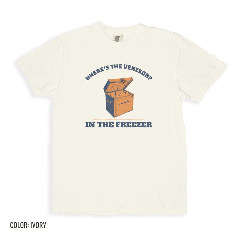 Ivory Comfort Colors t-shirt printed with the words Where'S The Venison? In The Freezer and a drawing of a vintage metal chest freezer with a pair of paper-wrapped butcher packages resting on top, folded flat lay