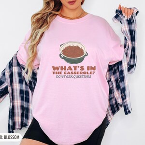 Blossom Comfort Colors t-shirt printed with the words What'S In The Casserole? Don'T Ask Questions and a drawing of a bubbly baked casserole in a rectangular ceramic baking dish topped with crispy fried oni, worn by a woman
