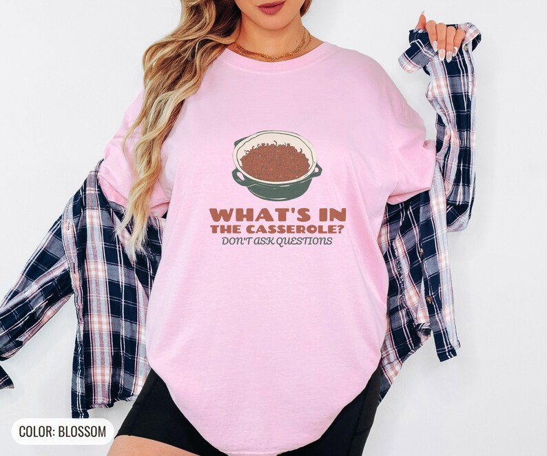 Blossom Comfort Colors t-shirt printed with the words What'S In The Casserole? Don'T Ask Questions and a drawing of a bubbly baked casserole in a rectangular ceramic baking dish topped with crispy fried oni, worn by a woman