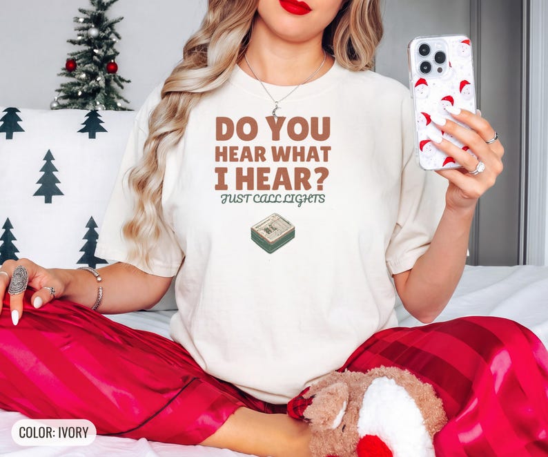 Ivory Comfort Colors t-shirt printed with the words Do You Hear What I Hear? Just Call Lights and a drawing of a rectangular electronic medical pager with a small holly sprig beside it, worn by a woman