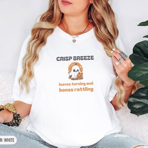 White Comfort Colors t-shirt printed with the words Crisp Breeze Leaves Turning And Bones Rattling and a drawing of a large cute smiling ghost beside a carved jack-o-lantern, worn by a woman