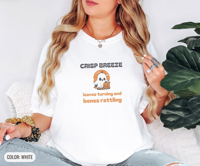 White Comfort Colors t-shirt printed with the words Crisp Breeze Leaves Turning And Bones Rattling and a drawing of a large cute smiling ghost beside a carved jack-o-lantern, worn by a woman
