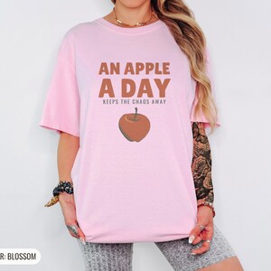 Blossom Comfort Colors t-shirt printed with the words An Apple A Day Keeps The Chaos Away and a drawing of a shiny red apple with a single green leaf and a tiny worm peeking out, worn by a woman