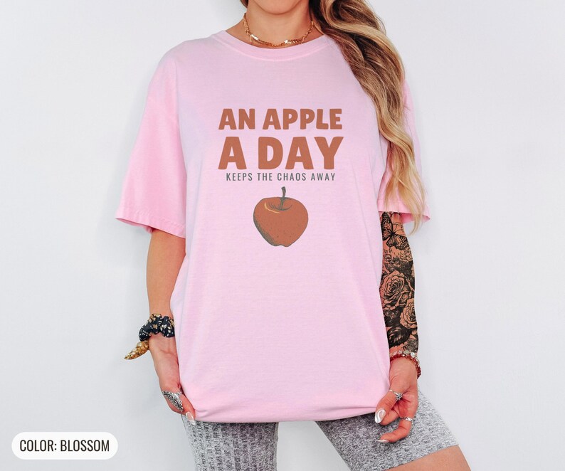 Blossom Comfort Colors t-shirt printed with the words An Apple A Day Keeps The Chaos Away and a drawing of a shiny red apple with a single green leaf and a tiny worm peeking out, worn by a woman