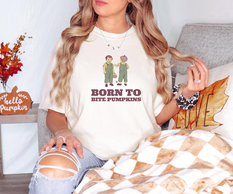 Ivory Comfort Colors t-shirt printed with the words Born To Bite Pumpkins and a drawing of pair of corduroy toddler overalls splattered with pumpkin pulp, worn by a woman
