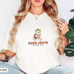 Ivory Comfort Colors t-shirt printed with the words Dear Santa The Ferret Did It and a drawing of a mischievous ferret dragging a striped Christmas stocking filled with shiny ornaments, worn by a woman
