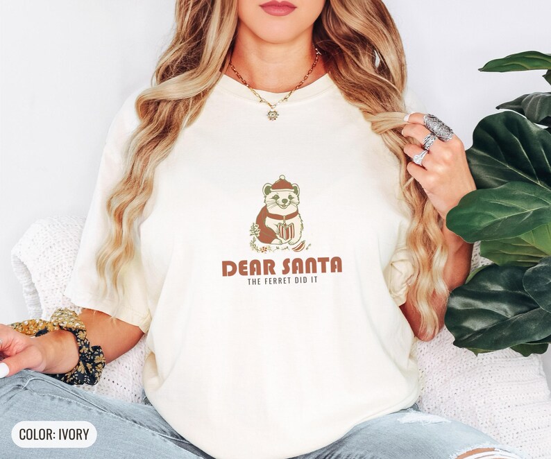 Ivory Comfort Colors t-shirt printed with the words Dear Santa The Ferret Did It and a drawing of a mischievous ferret dragging a striped Christmas stocking filled with shiny ornaments, worn by a woman