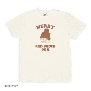Ivory Comfort Colors t-shirt printed with the words Merry And Under Par and a drawing of a dimpled golf ball wearing a tiny red and white striped knit winter beanie with a green p, folded flat lay