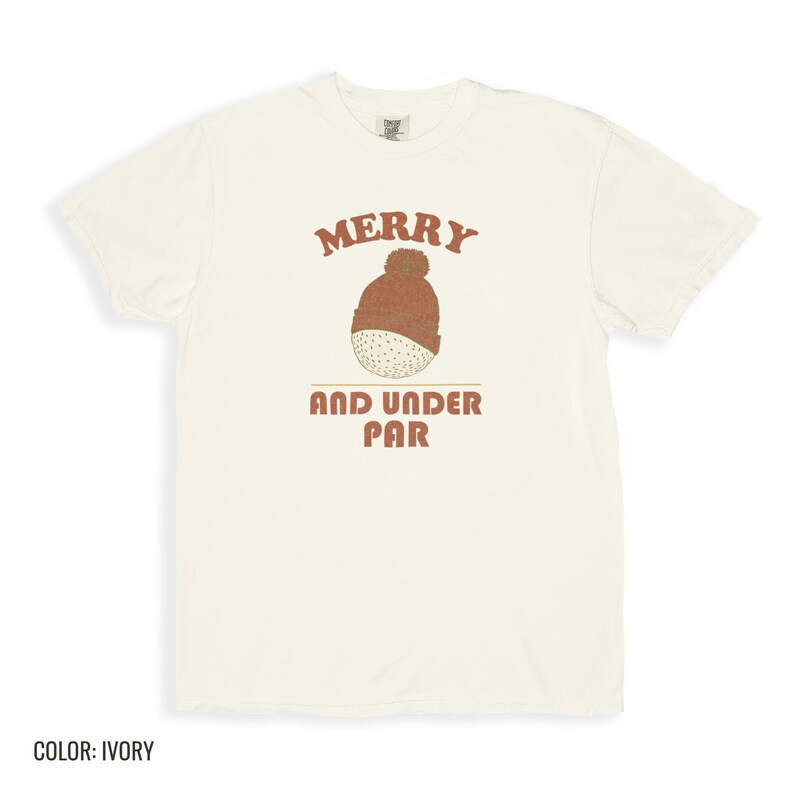 Ivory Comfort Colors t-shirt printed with the words Merry And Under Par and a drawing of a dimpled golf ball wearing a tiny red and white striped knit winter beanie with a green p, folded flat lay