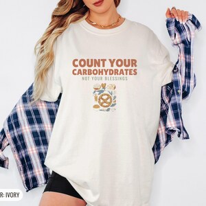 Ivory Comfort Colors t-shirt printed with the words Count Your Carbohydrates Not Your Blessings and a drawing of a golden-brown braided pretzel with coarse salt grains, worn by a woman