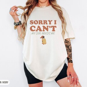 Ivory Comfort Colors t-shirt printed with the words Sorry I Can'T My Dog Needs Me and a drawing of a sleeping dachshund wearing a patterned knit sweater, worn by a woman
