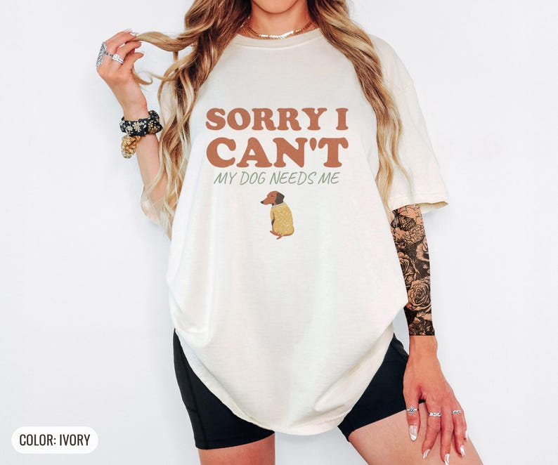 Ivory Comfort Colors t-shirt printed with the words Sorry I Can'T My Dog Needs Me and a drawing of a sleeping dachshund wearing a patterned knit sweater, worn by a woman