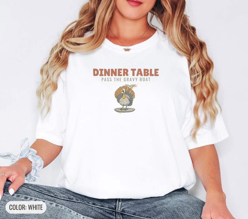 White Comfort Colors t-shirt printed with the words Dinner Table Pass The Gravy Boat and a drawing of a golden roast turkey on a platter, worn by a woman