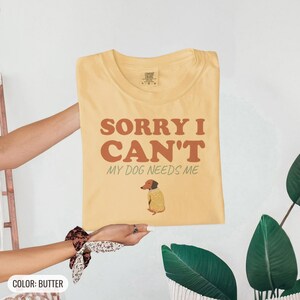 Butter Comfort Colors t-shirt printed with the words Sorry I Can'T My Dog Needs Me and a drawing of a sleeping dachshund wearing a patterned knit sweater, folded flat lay