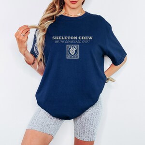 True Navy Comfort Colors t-shirt printed with the words Skeleton Crew On The Graveyard Shift and a drawing of a detailed medical defibrillator with its paddle cords forming a heart shape, flanked by t, worn by a woman