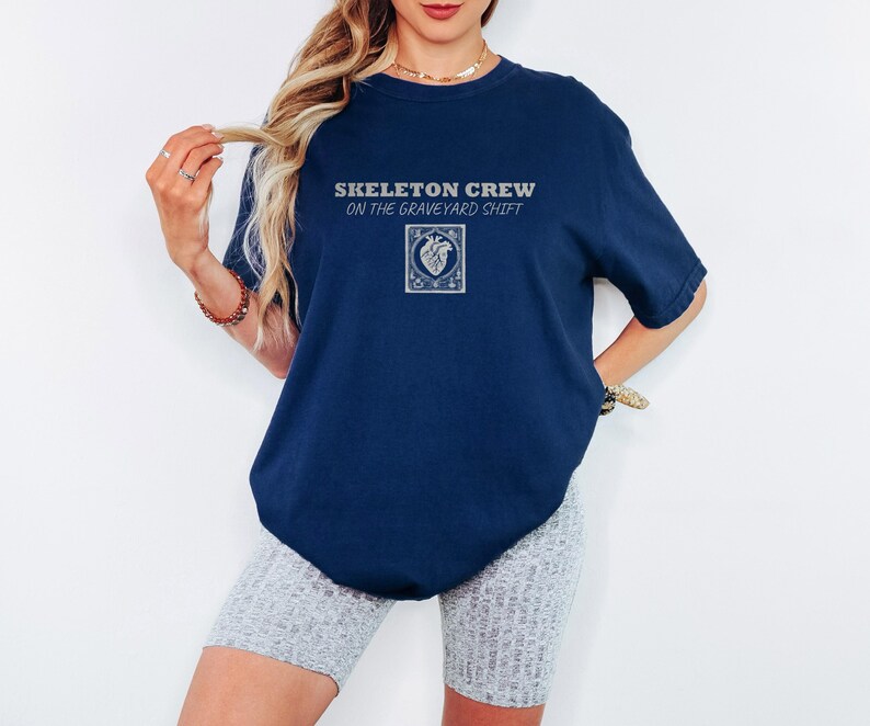 True Navy Comfort Colors t-shirt printed with the words Skeleton Crew On The Graveyard Shift and a drawing of a detailed medical defibrillator with its paddle cords forming a heart shape, flanked by t, worn by a woman