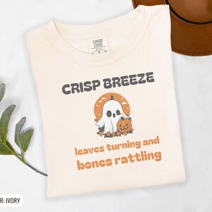 Ivory Comfort Colors t-shirt printed with the words Crisp Breeze Leaves Turning And Bones Rattling and a drawing of a large cute smiling ghost beside a carved jack-o-lantern, folded flat lay