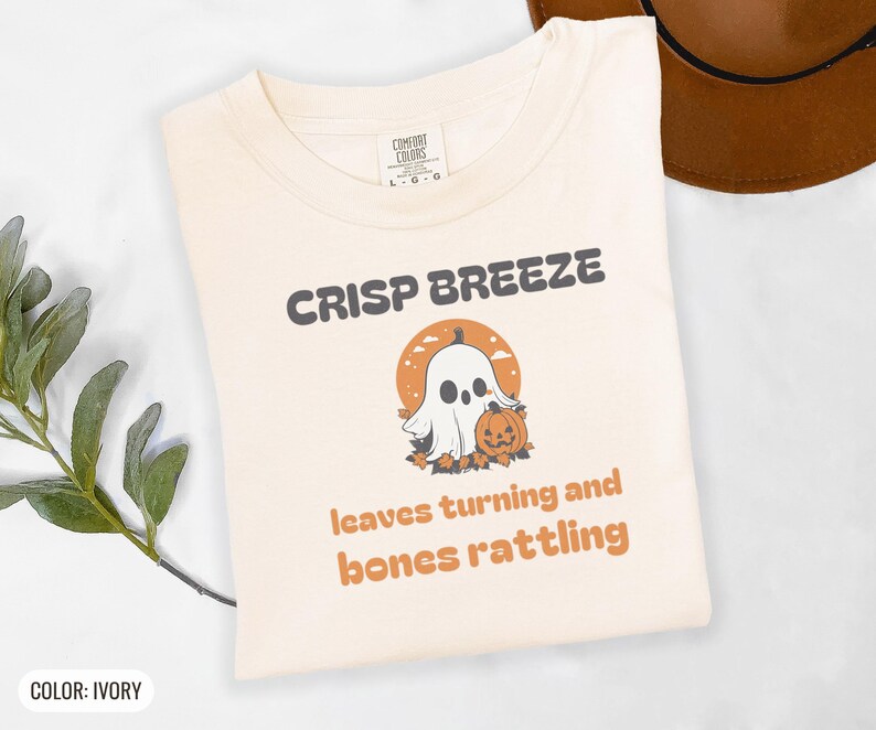 Ivory Comfort Colors t-shirt printed with the words Crisp Breeze Leaves Turning And Bones Rattling and a drawing of a large cute smiling ghost beside a carved jack-o-lantern, folded flat lay