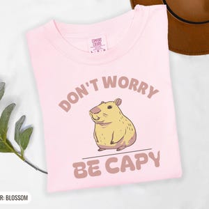 Blossom Comfort Colors t-shirt printed with the words Don'T Worry Be Capy and a drawing of a happy capybara with a small orange citrus slice on its head beside a green lily pad, folded flat lay