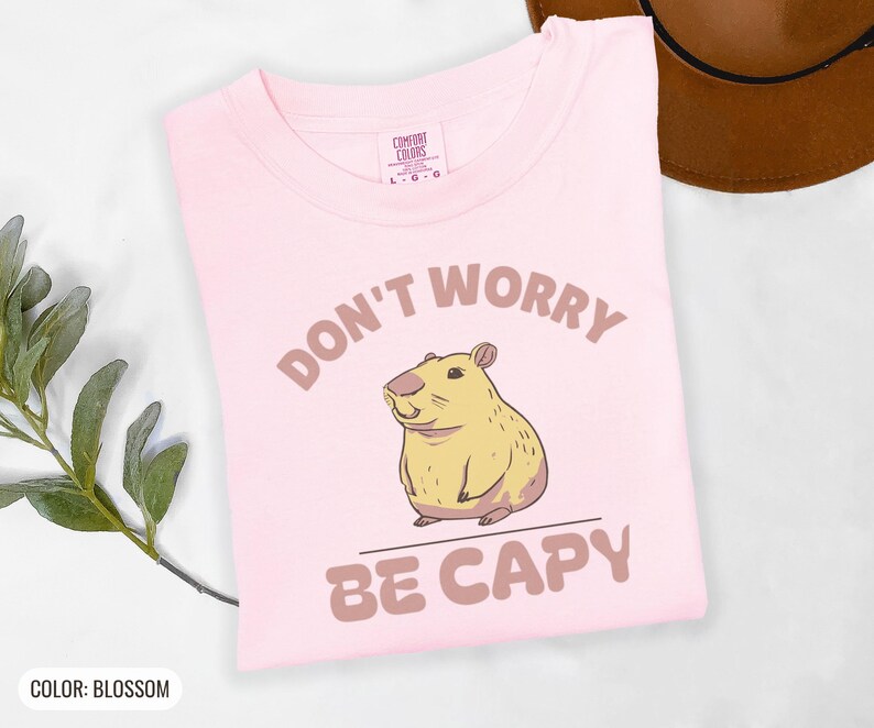Blossom Comfort Colors t-shirt printed with the words Don'T Worry Be Capy and a drawing of a happy capybara with a small orange citrus slice on its head beside a green lily pad, folded flat lay