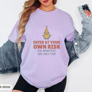 Orchid Comfort Colors t-shirt printed with the words Enter At Your Own Risk The Monsters Are Only Five and a drawing of a cute, smiling little green monster wearing a tiny paper party hat and holding a crayon, worn by a woman