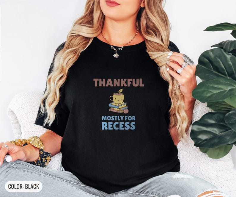 Black Comfort Colors t-shirt printed with the words Thankful Mostly For Recess and a drawing of a stack of retro school books topped with a glossy red apple and a steaming ceramic coffee, worn by a woman