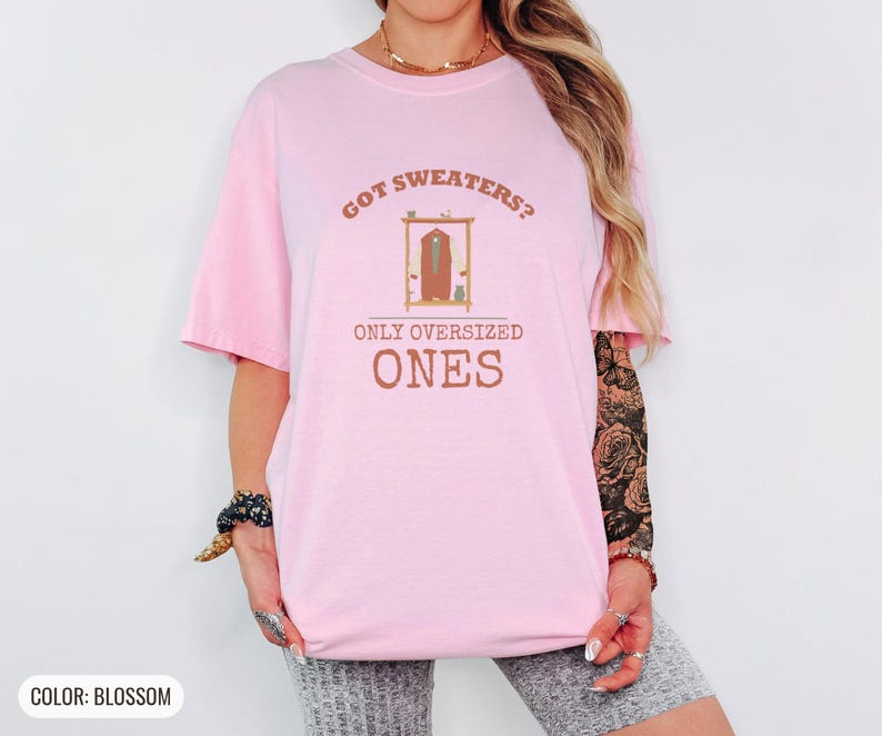 Blossom Comfort Colors t-shirt printed with the words Got Sweaters? Only Oversized Ones and a drawing of a knitted wool cardigan hanging from a simple wooden clothes hanger, worn by a woman