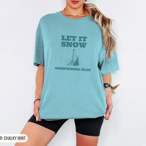 Chalky Mint Comfort Colors t-shirt printed with the words Let It Snow Somewhere Else and a drawing of a metal snow shovel propped beside an empty bag of rock salt, worn by a woman