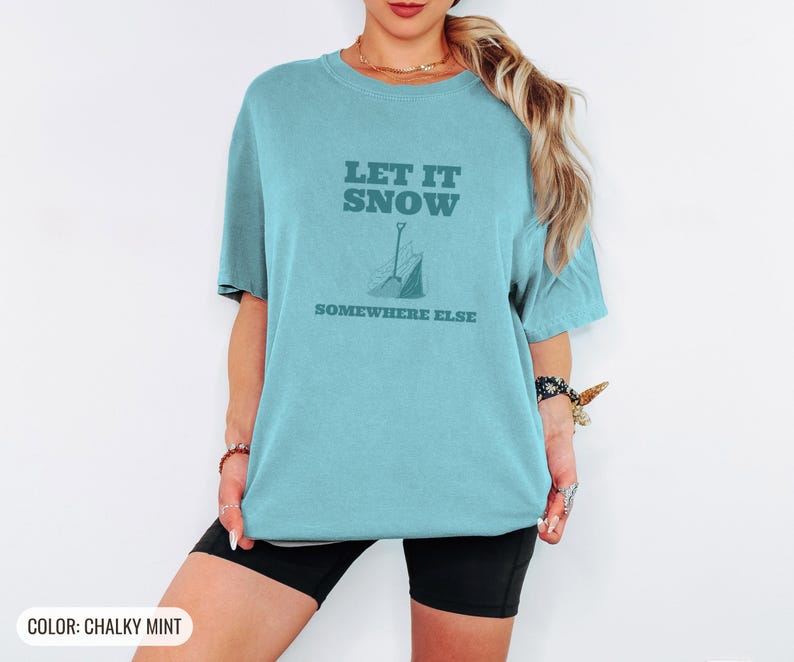 Chalky Mint Comfort Colors t-shirt printed with the words Let It Snow Somewhere Else and a drawing of a metal snow shovel propped beside an empty bag of rock salt, worn by a woman