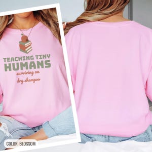 Blossom Comfort Colors t-shirt printed with the words Teaching Tiny Humans Surviving On Dry Shampoo and a drawing of a small neat stack of three closed hardcover books with PLAIN blank solid-colour spines an, worn by a woman