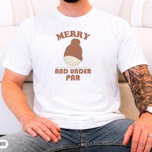 White Comfort Colors t-shirt printed with the words Merry And Under Par and a drawing of a dimpled golf ball wearing a tiny red and white striped knit winter beanie with a green p, worn by a man