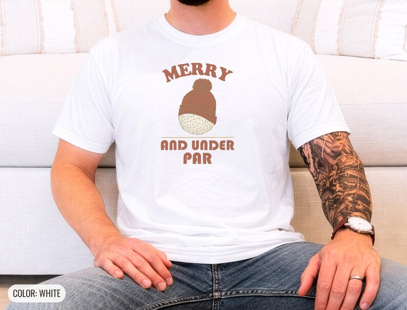 White Comfort Colors t-shirt printed with the words Merry And Under Par and a drawing of a dimpled golf ball wearing a tiny red and white striped knit winter beanie with a green p, worn by a man
