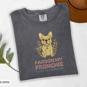 Pepper Comfort Colors t-shirt printed with the words Pardon My Frenchie Under The Mistletoe and a drawing of a vintage brass French Bulldog bottle opener lying next to a sprig of red-berried mistleto, folded flat lay