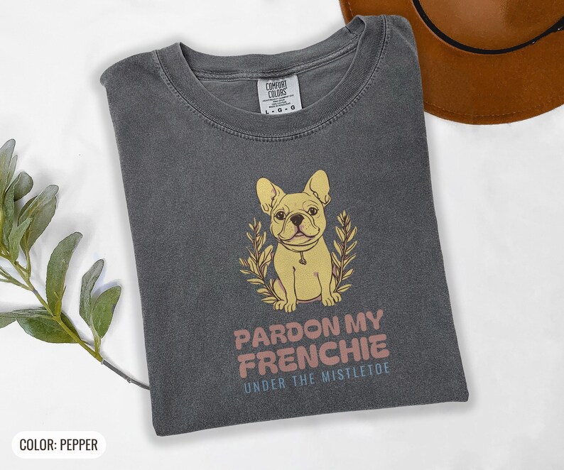 Pepper Comfort Colors t-shirt printed with the words Pardon My Frenchie Under The Mistletoe and a drawing of a vintage brass French Bulldog bottle opener lying next to a sprig of red-berried mistleto, folded flat lay