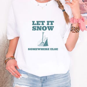 White Comfort Colors t-shirt printed with the words Let It Snow Somewhere Else and a drawing of a metal snow shovel propped beside an empty bag of rock salt, worn by a woman