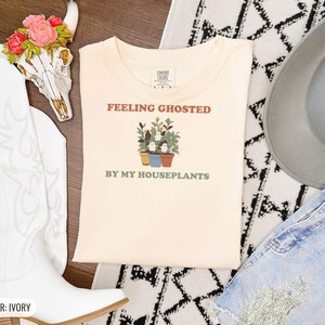 Ivory Comfort Colors t-shirt printed with the words Feeling Ghosted By My Houseplants and a drawing of a compact, self-contained group of three small potted houseplants with tiny, friendly whit, folded flat lay