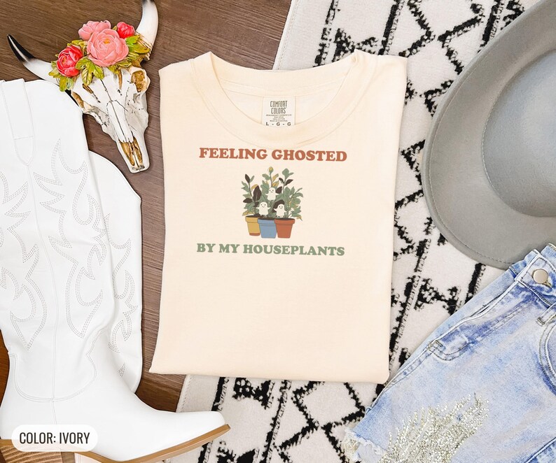 Ivory Comfort Colors t-shirt printed with the words Feeling Ghosted By My Houseplants and a drawing of a compact, self-contained group of three small potted houseplants with tiny, friendly whit, folded flat lay